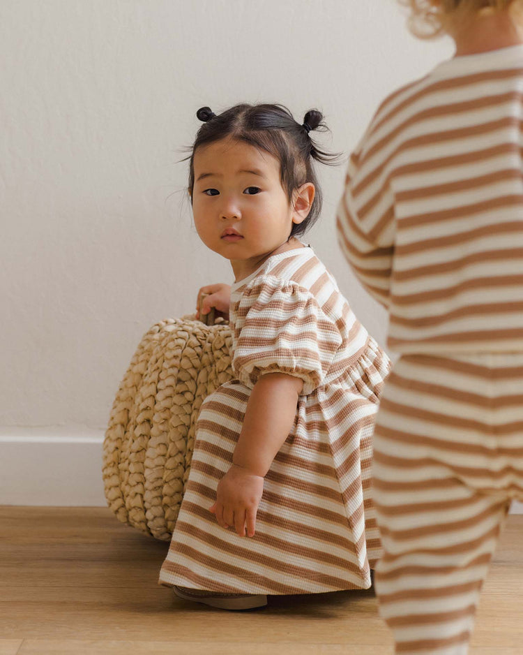 A young child wearing the quincy mae darla dress in spice stripe, made from organic cotton, kneels by a woven basket and looks toward the camera. Another child stands nearby with their back turned, also dressed in coordinating spice stripe clothing.