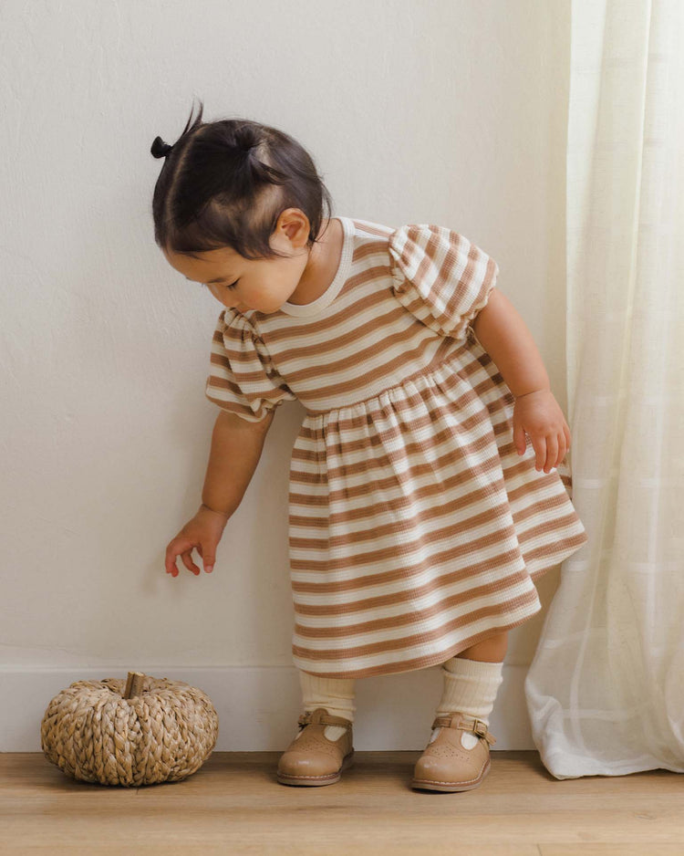 A toddler wearing the quincy mae darla dress in spice stripe and tan shoes looks down at a woven pumpkin decoration on the floor, standing near a cream-colored curtain.