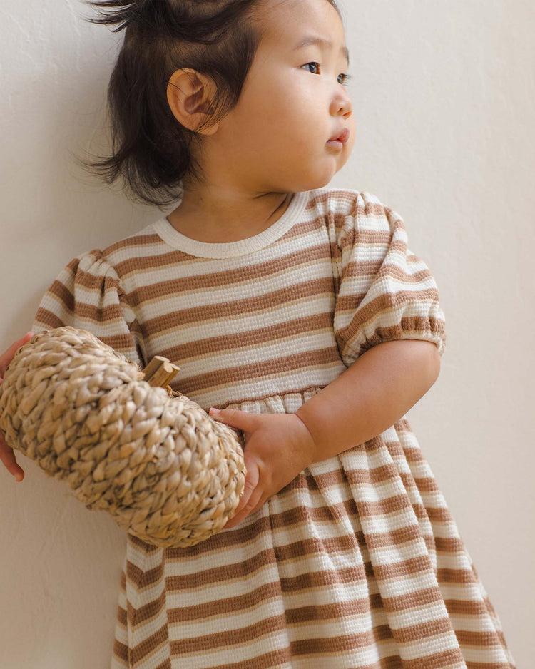 A toddler wears the quincy mae darla dress in spice stripe—an organic cotton piece in brown and white stripes—while holding a round, woven basket and looking to the side against a light background.