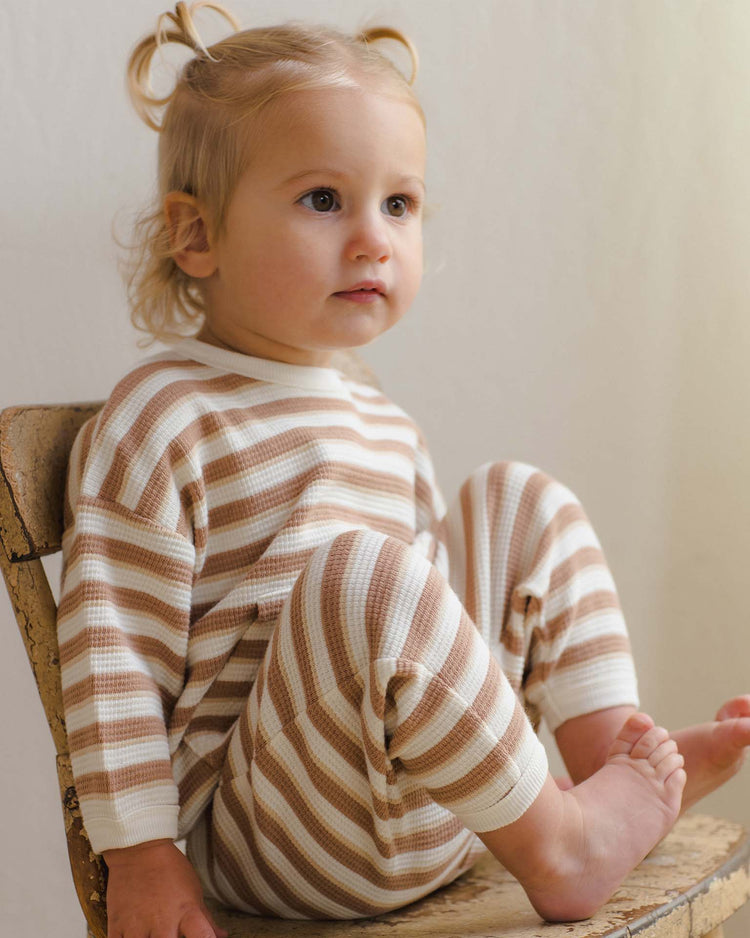 A young child with light hair sits sideways on a rustic wooden chair, wearing the quincy mae waffle slouch set in spice stripe—soft organic cotton pajamas in beige and white—looking slightly off camera with a calm expression.