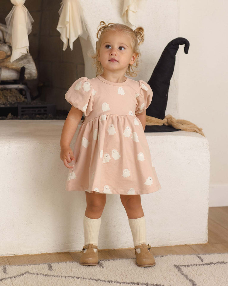 A toddler wearing the quincy mae darla dress in sweet ghost—crafted from organic cotton—stands indoors, paired with tan shoes and cream socks. Behind her is a white fireplace decorated for Halloween with a black witch hat and cream-colored fabric.