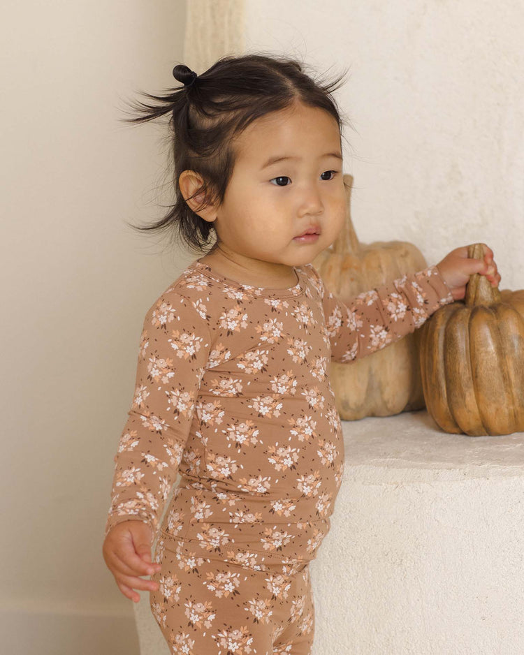 A young child wears the quincy mae bamboo pajama set in fall floral, standing indoors while holding a decorative pumpkin. Two pumpkins rest on a white surface in the background.
