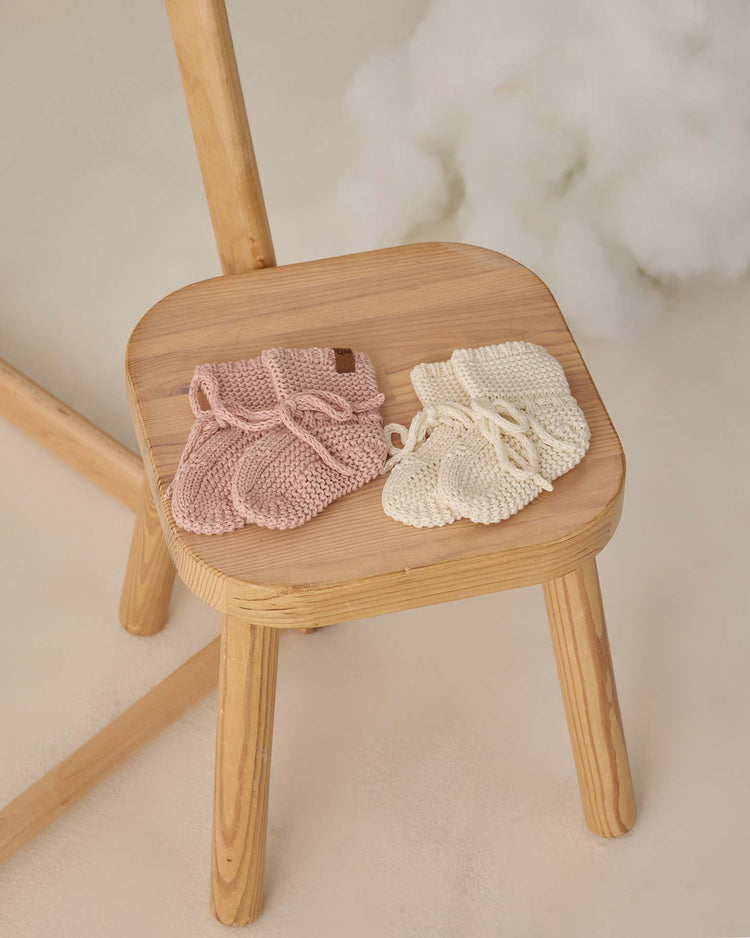 A wooden stool showcases Quincy Mae knit booties in blush, crafted from organic cotton. The background is light with soft, fluffy decor.