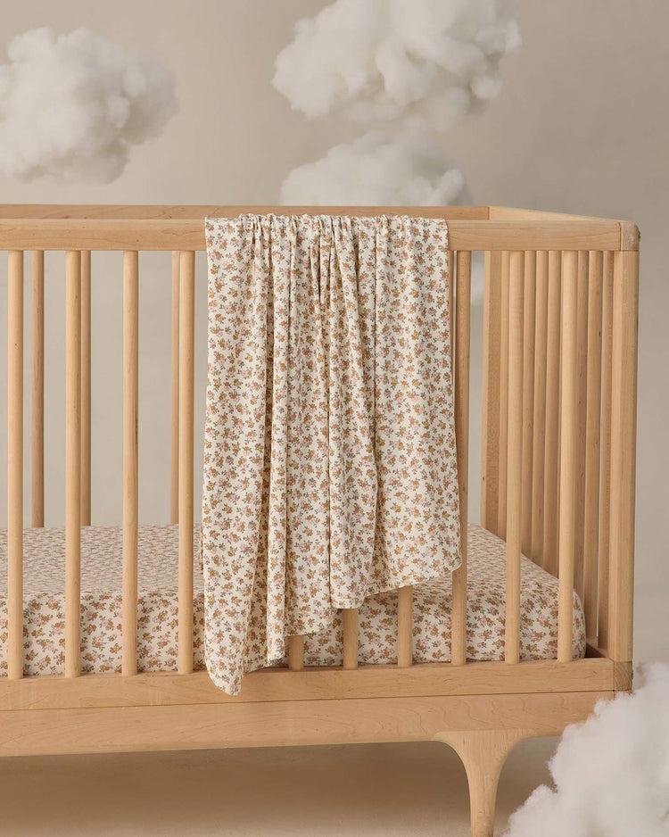 A bamboo swaddle in wildflower by quincy mae is draped over a wooden crib with a matching sheet, while fluffy clouds hang against a beige background for a dreamy look.