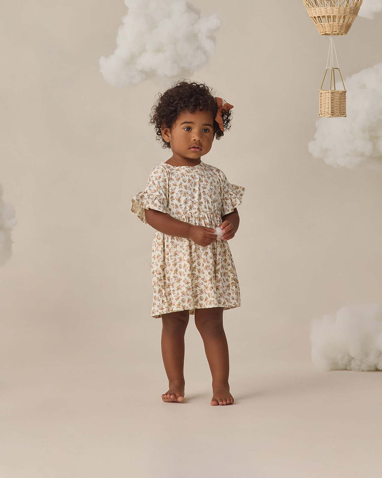 A young child with curly hair and a bow stands barefoot, wearing the quincy mae bamboo babydoll dress in wildflower. Fluffy clouds and a wicker hot air balloon set a dreamy, whimsical background.