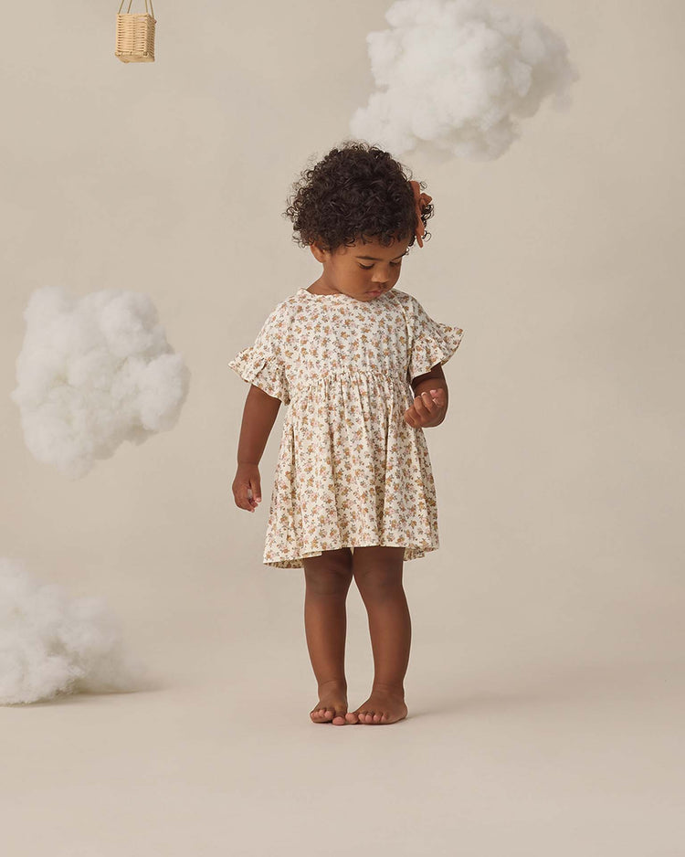 A young child with curly hair stands barefoot, looking down, wearing the quincy mae bamboo babydoll dress in wildflower. The background is beige with fluffy white clouds hanging as decoration.