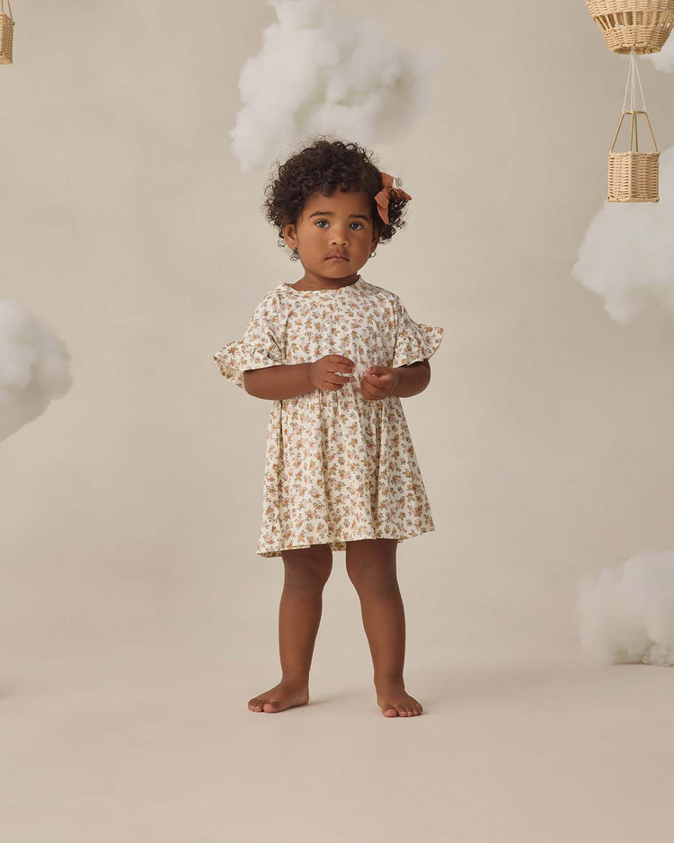 A young child with curly hair stands barefoot against a beige background, wearing the quincy mae bamboo babydoll dress in wildflower with ruffle sleeves and a matching bow in her hair.