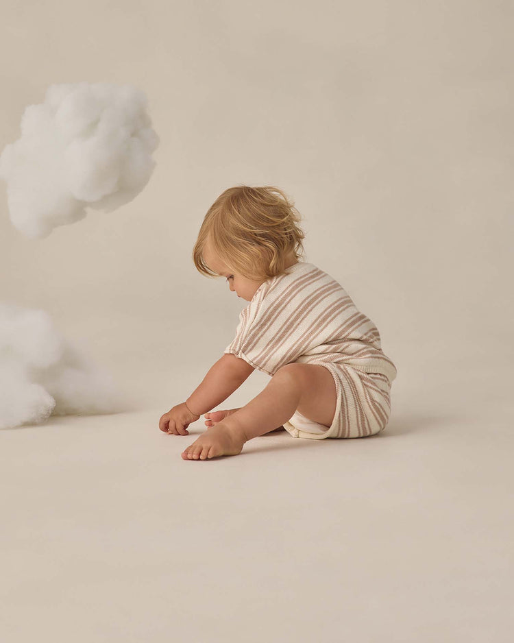 A young child with light brown hair wears the quincy mae relaxed summer knit set in oat stripe, made from organic cotton, sitting on the floor near a fluffy white cloud decoration against a neutral beige background.