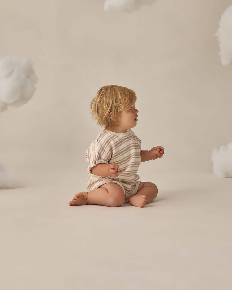 A young child with blonde hair sits barefoot on a neutral background, wearing the quincy mae relaxed summer knit set in oat stripe, surrounded by fluffy, cloud-like decorations for a dreamy touch.