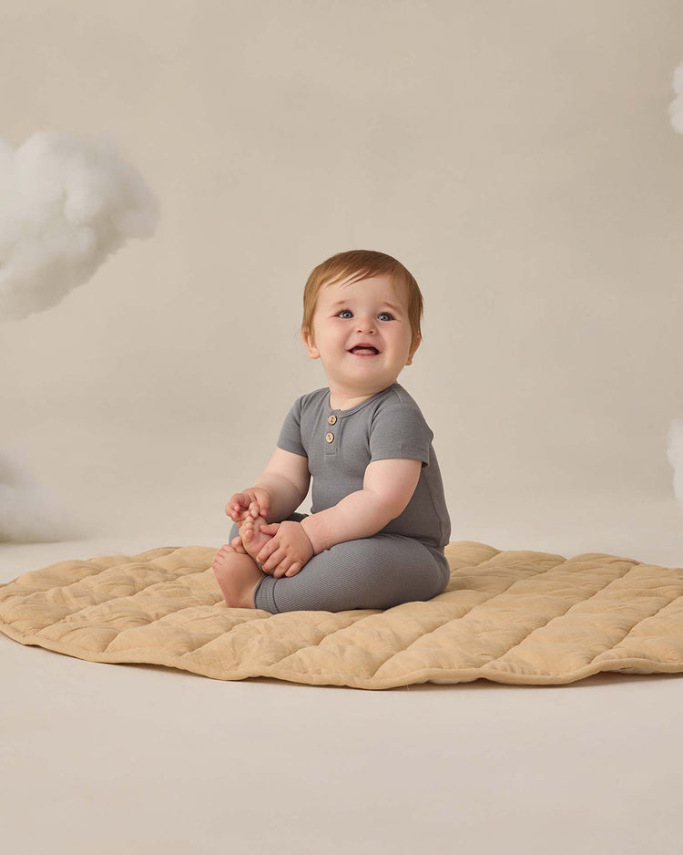 A smiling baby wearing quincy mae's ribbed legging in ocean and a gray outfit sits on a beige quilted mat, holding their feet against a light background with soft, fluffy clouds.