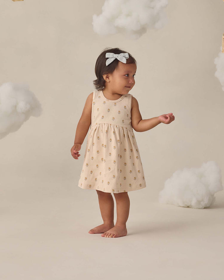 A young child in the quincy mae ribbed tank dress in buttercup stands barefoot on a light background, surrounded by fluffy cloud props, with a flower print and matching bloomers, wearing a white bow headband.
