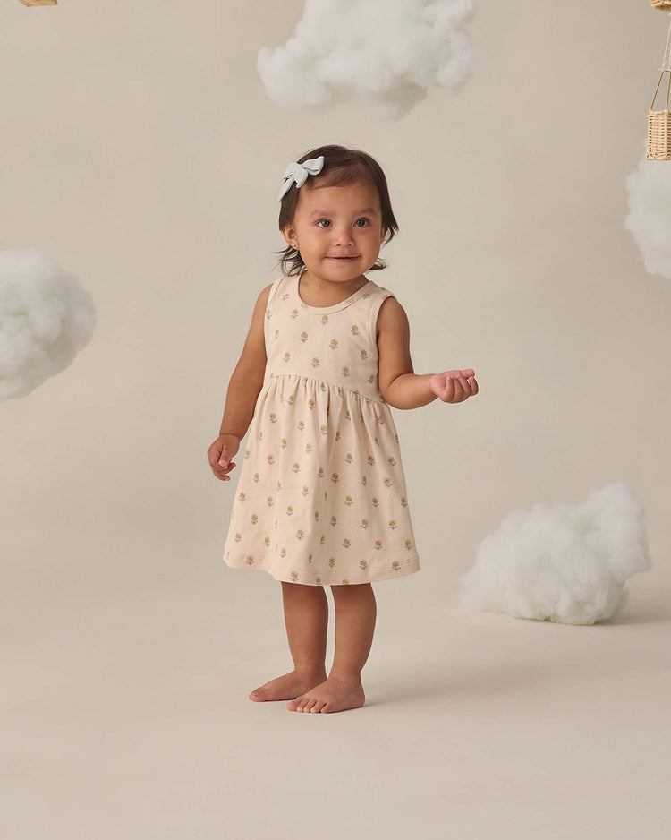 A young girl stands barefoot on a beige floor, wearing the quincy mae ribbed tank dress in buttercup with organic cotton bloomers peeking out and a white bow in her hair, surrounded by fluffy, cloud-like props.