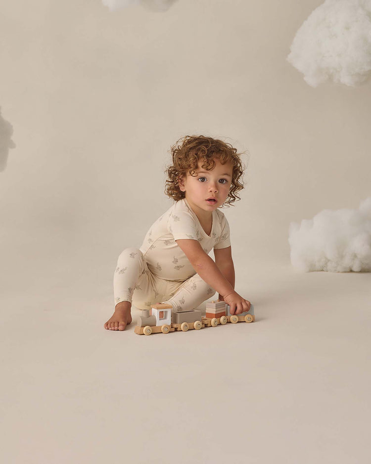 A young child with curly hair sits barefoot, wearing the quincy mae bamboo short sleeve pajama set in elephants, and plays with a wooden toy train. Soft clouds float in the beige background, creating a dreamy scene.
