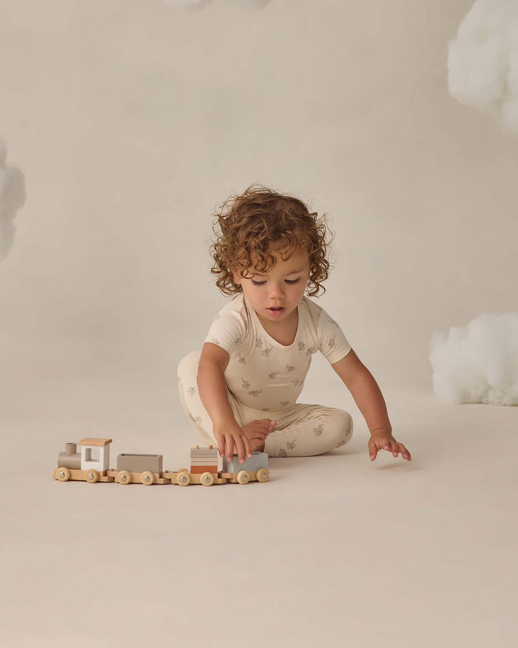 A young child with curly hair, dressed in the quincy mae bamboo short sleeve pajama set in elephants, sits on the floor playing with a wooden toy train. The background features neutral tones and fluffy cloud-like decorations.
