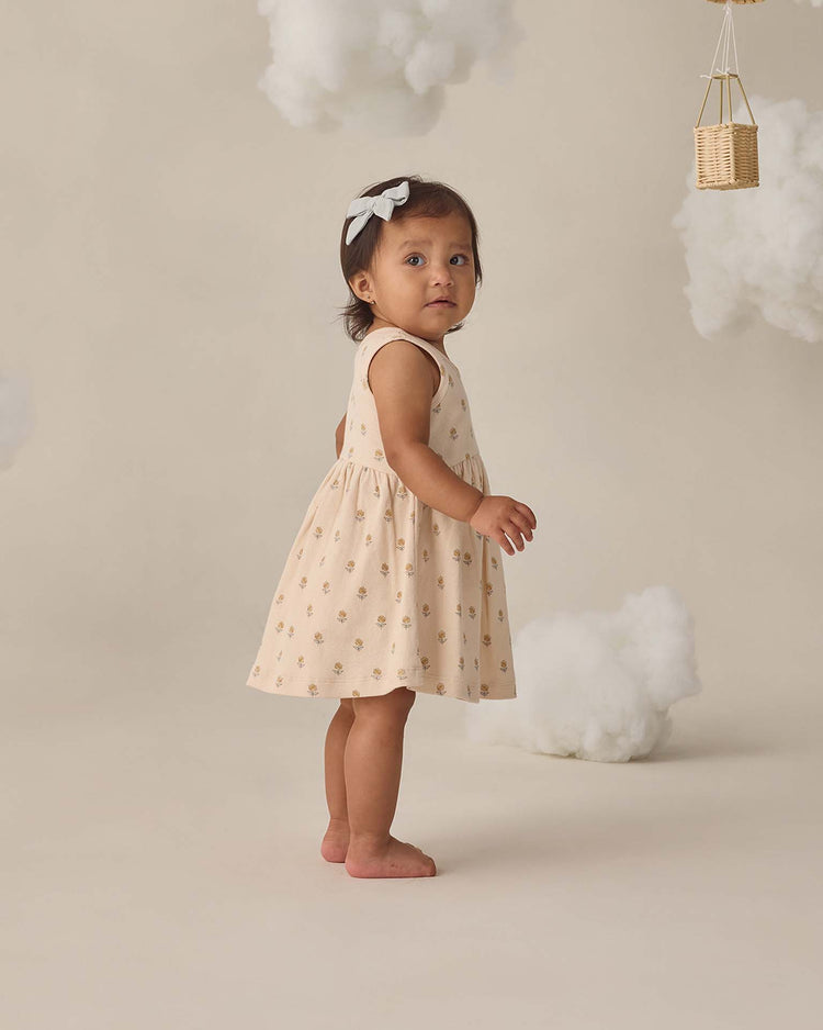 A young child with a white bow in her hair wears the quincy mae ribbed tank dress in buttercup as she stands barefoot on a neutral set decorated with fluffy white clouds and a small hanging basket.