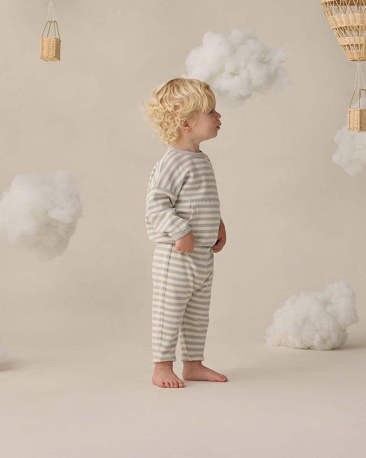 A young child with curly blonde hair stands barefoot in profile, wearing the quincy mae waffle sweater set in sky stripe. Fluffy clouds and hanging basket props create a dreamy background.