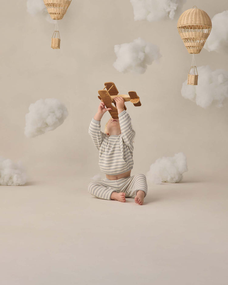 A baby in the quincy mae waffle sweater set in sky stripe sits on the floor, holding a wooden toy airplane. Fluffy artificial clouds and wicker hot air balloons surround them, creating a whimsical, dreamy scene.