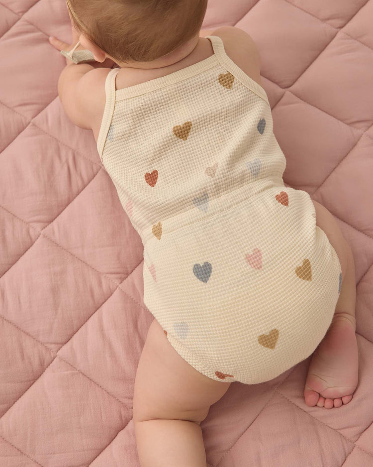 A baby lies on a pink quilted blanket, wearing the quincy mae waffle cinch romper in confetti hearts—a cream-colored organic cotton outfit with small, multicolored hearts—viewed from above.