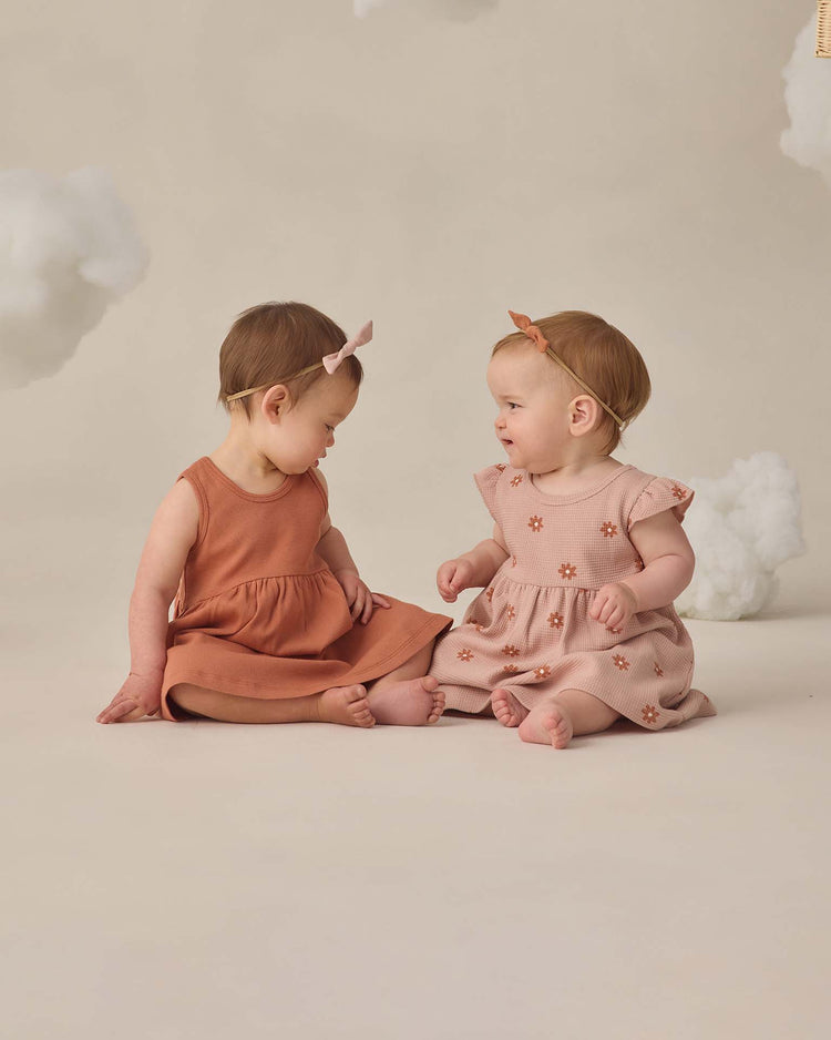Two babies sit on the floor against a beige background with soft white clouds, wearing quincy mae ribbed tank dresses in poppy and matching bows, paired with bloomers. They smile sweetly at each other.
