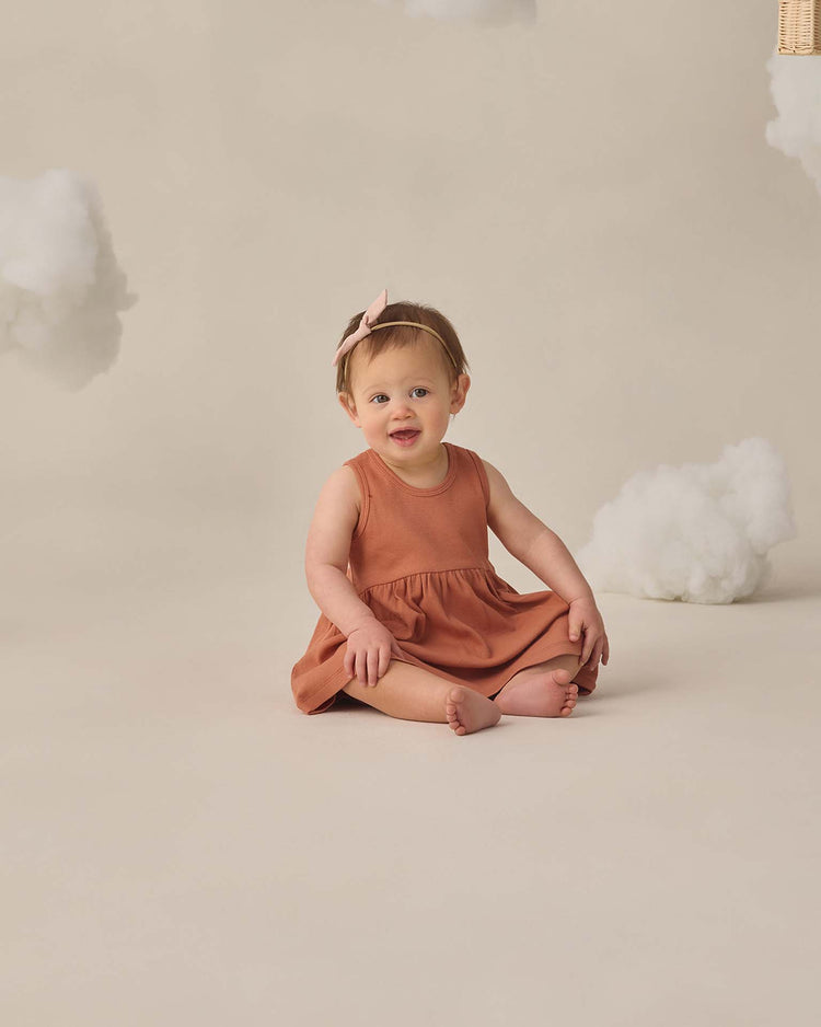A smiling baby sits on the floor against a beige background with fluffy white cloud props, wearing the quincy mae ribbed tank dress in poppy with matching bloomers and a headband.