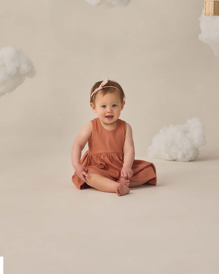 A baby in the quincy mae ribbed tank dress in poppy and a pink headband sits barefoot on a cream floor, smiling, with fluffy white clouds in the background for a whimsical touch.