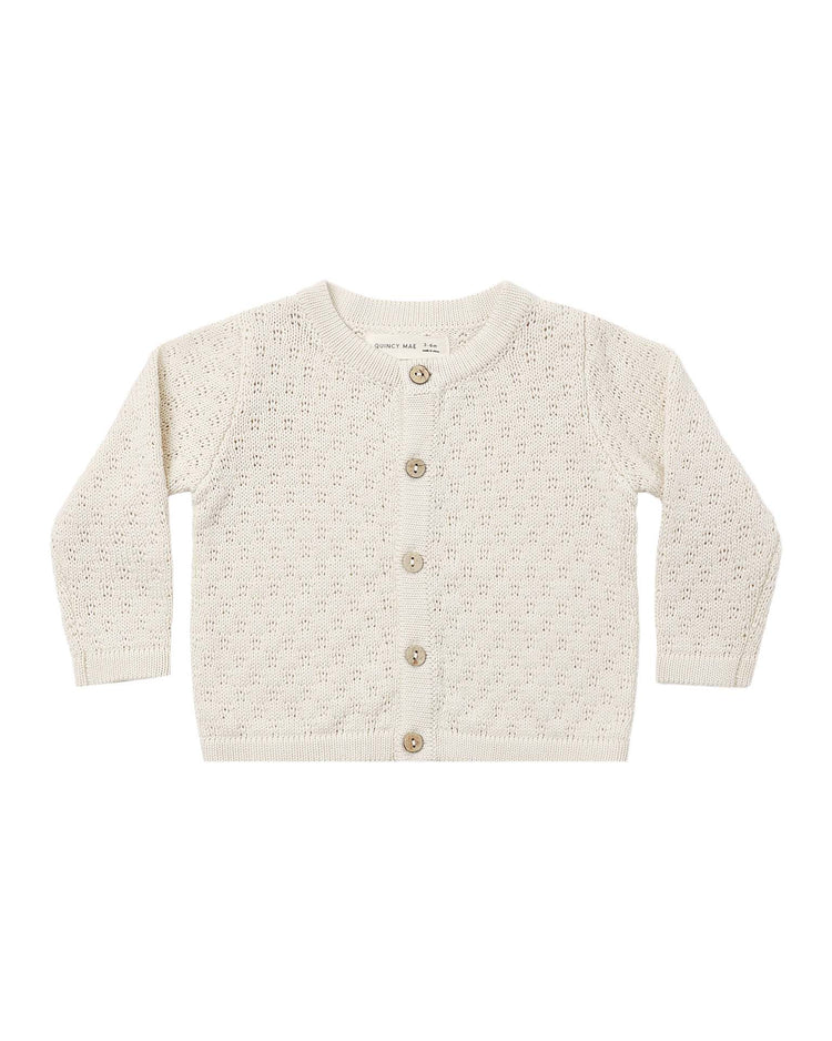 The "knit cardigan in natural" by quincy mae is a cream-colored, long-sleeve baby cardigan featuring a delicate textured knit pattern and five charming coconut buttons down the front, shown laid flat on a white background.