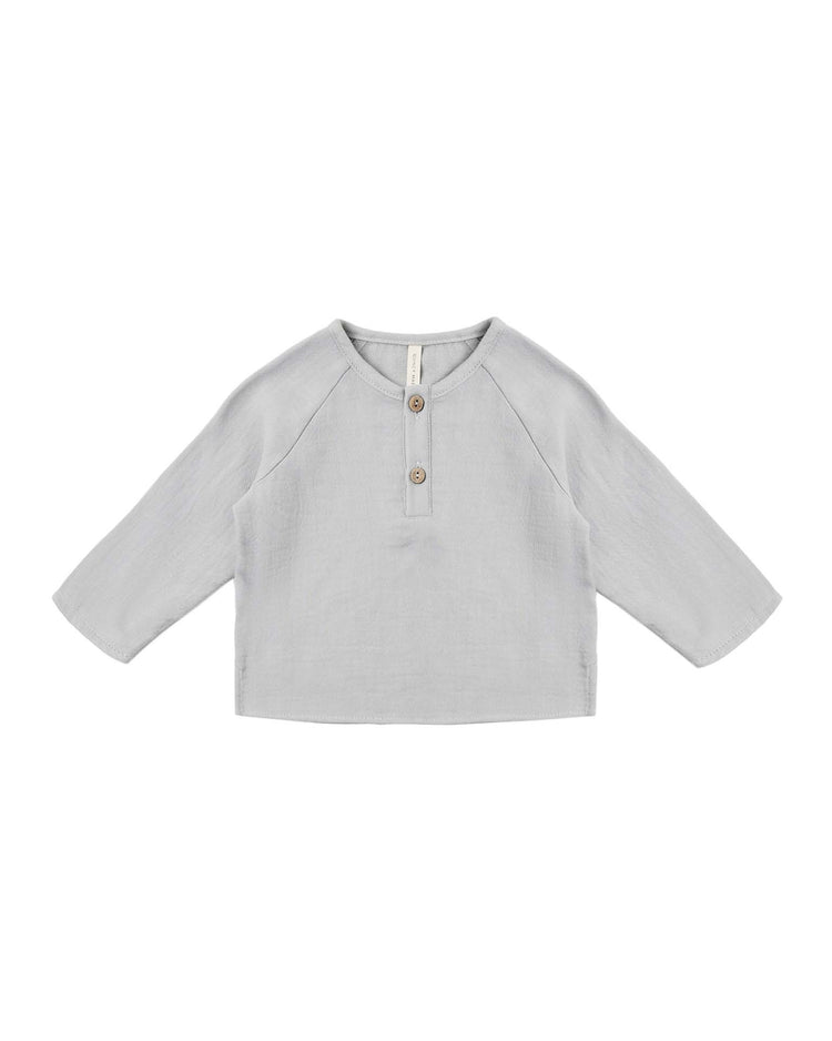 The zion shirt in cloud by quincy mae is a light gray, long-sleeve organic cotton baby shirt with a relaxed, boxy fit, featuring raglan sleeves and two brown buttons on the front placket, shown laid flat against a white background.