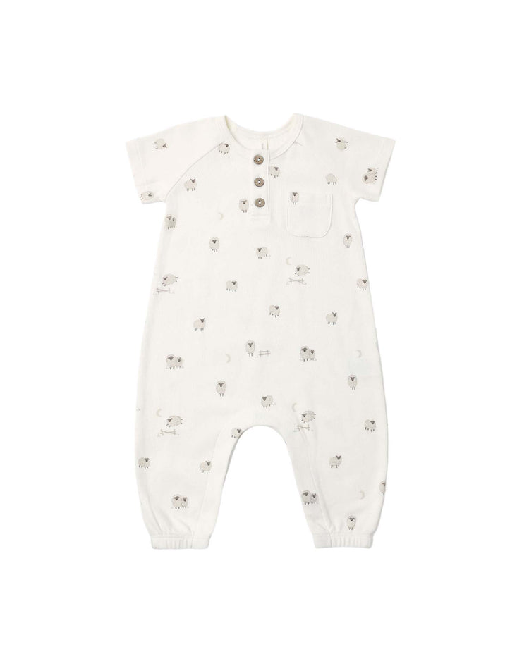 The "short sleeve jumpsuit in sheep" by quincy mae is a white baby romper crafted from soft organic cotton, featuring short sleeves, three chest buttons, a small chest pocket, and an allover subtle print of gray sheep and moons. Designed with elastic cuffs at the legs for added comfort, this adorable outfit is perfect for little ones.
