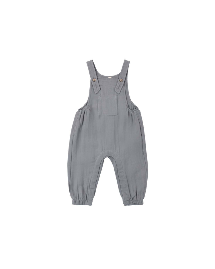The baby overall in slate by quincy mae is a gray, sleeveless organic cotton design featuring adjustable shoulder straps, a front pocket, and coconut button closures. The ankles are gathered for a comfortable fit, and the soft, lightweight fabric is displayed laid flat on a white background.