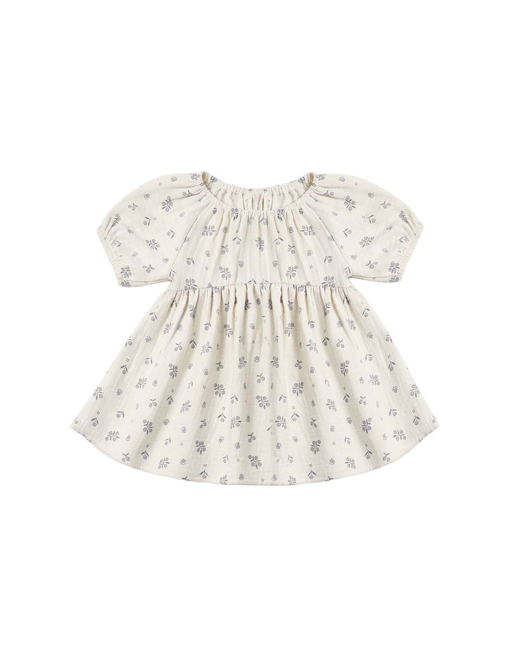 The "bella dress in winter berry" by Quincy Mae is an organic cotton baby girl dress in a light cream color, featuring short puff sleeves, a gathered skirt, and a subtle winter berry print in soft grayish-purple tones.