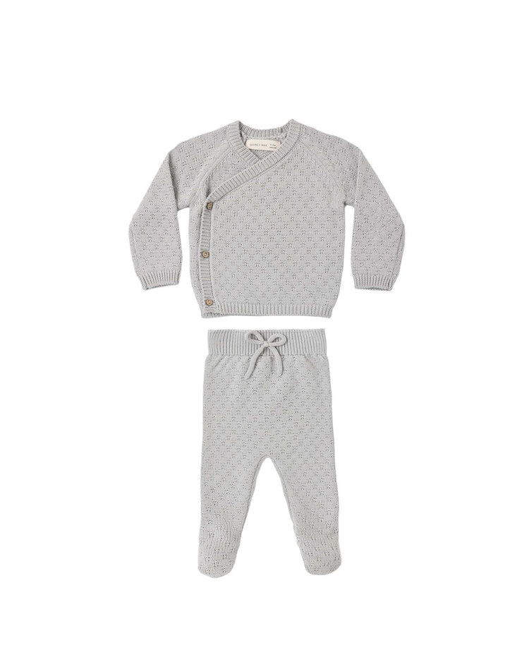 The "knit wrap top + pant set in cloud" by quincy mae is a light gray baby outfit made from soft organic cotton. The set includes a wrap-style long-sleeve top with side buttons and matching pants with a drawstring waist, both featuring a subtle dotted pattern for extra charm. This cozy newborn ensemble is displayed laid flat on a white background.