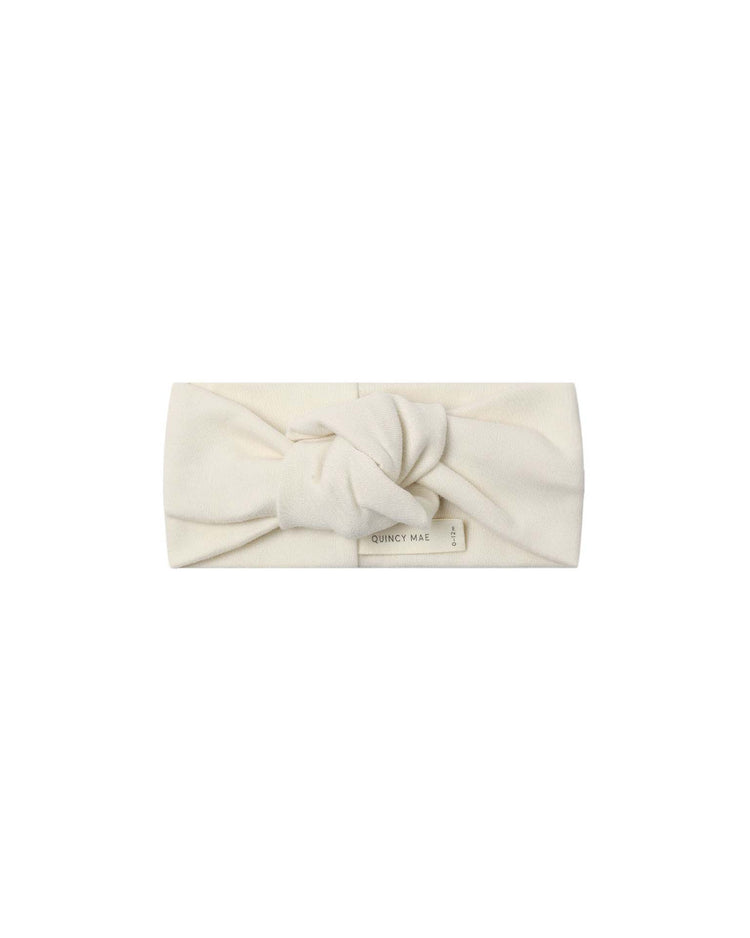 The knotted headband in natural by quincy mae is a soft, cream-colored baby accessory made from organic cotton, featuring a central knot and a small “QUINCY MAE” label on the front. It is displayed against a plain white background.