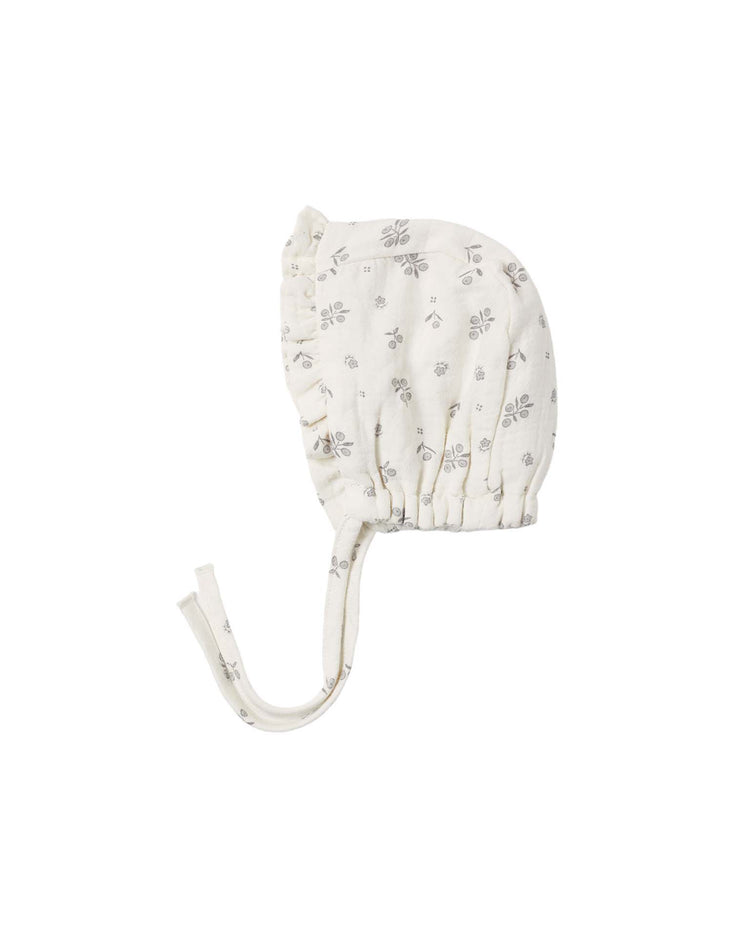 A white "woven ruffle bonnet in winter berry" by quincy mae, crafted from organic cotton with delicate gray floral designs and two fabric ties, is displayed on a plain white background.