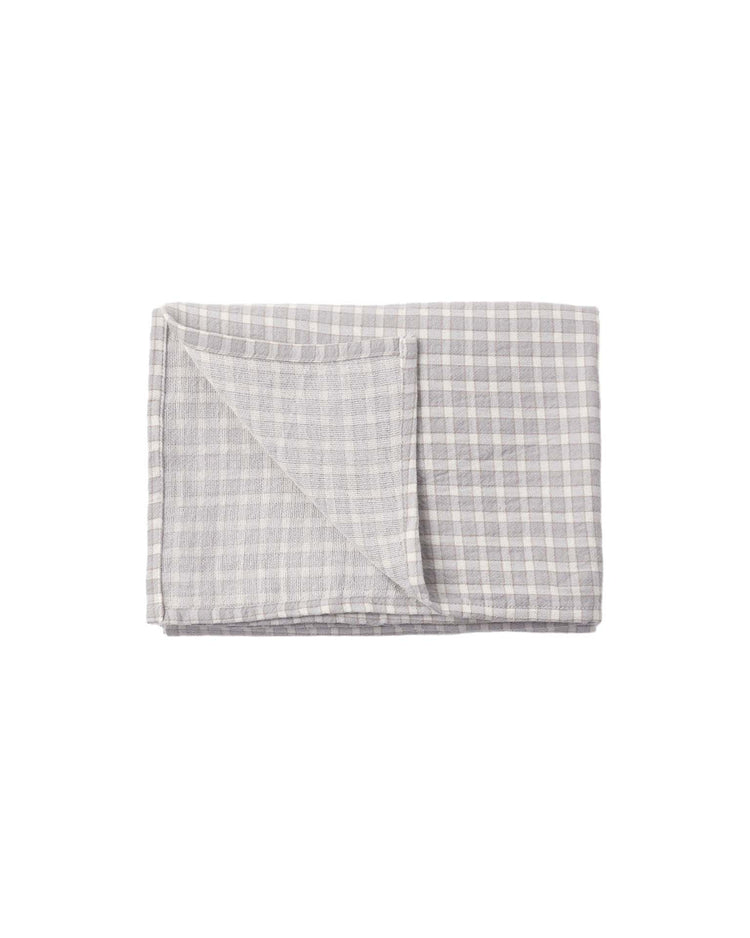A folded, light blue gingham woven swaddle from quincy mae is displayed on a plain white background. One corner is turned up to reveal the reverse side, evoking the timeless look of classic kitchen linens.