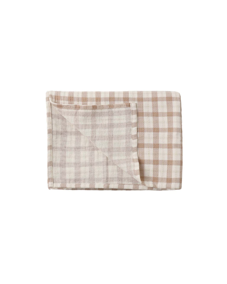 A folded quincy mae woven swaddle in rustic plaid, made from soft organic cotton, with one corner partially turned over to reveal the beige and white gingham-patterned reverse side.