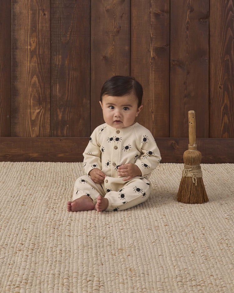 A baby sits on a beige textured rug, wearing the quincy mae waffle long sleeve jumpsuit in bitsy spider—an organic cotton outfit in cream with a playful spider print. A small broom stands upright beside the baby against a wooden wall backdrop.