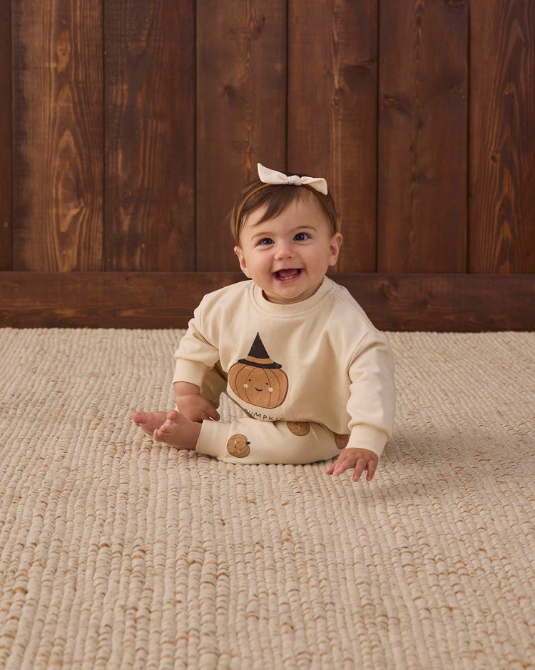 A smiling baby with brown hair and a bow sits on a textured beige rug, wearing the Quincy Mae relaxed fleece sweatshirt in lil pumpkin, in front of a wooden wall.