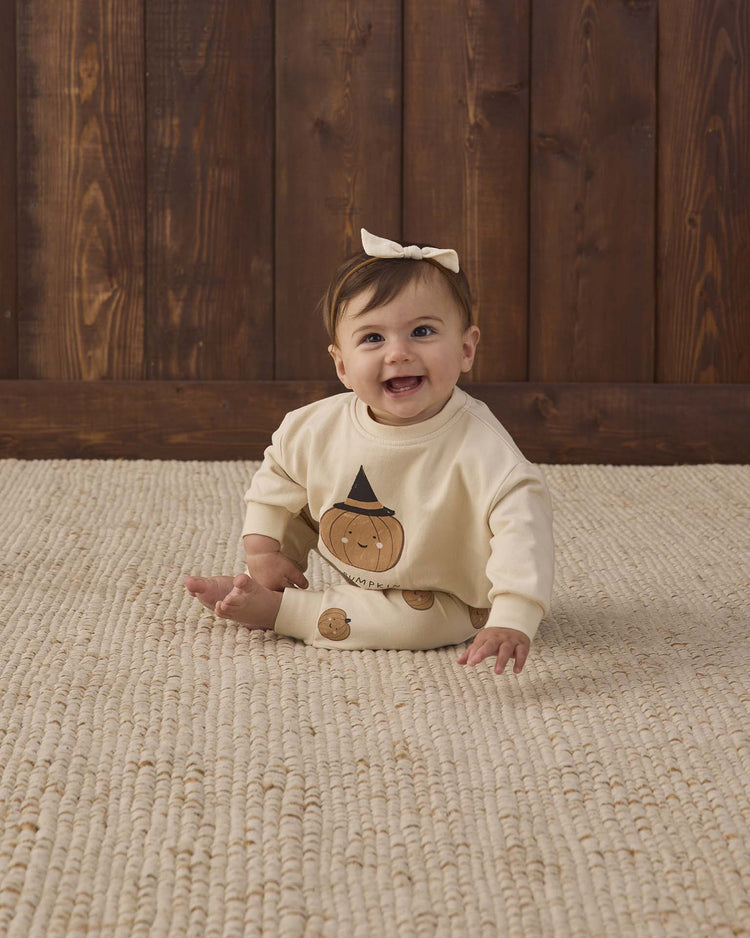 A smiling baby with brown hair and a bow sits on a textured beige rug, wearing the Quincy Mae relaxed fleece sweatshirt in lil pumpkin, in front of a wooden wall.