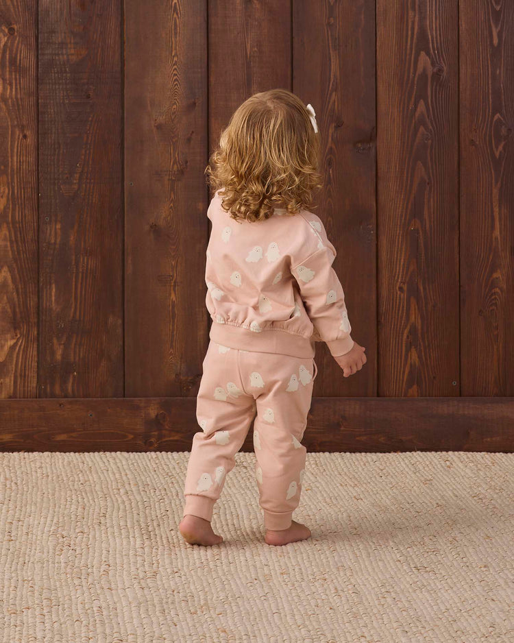 A young child with curly hair, wearing the quincy mae relaxed fleece sweatpant in sweet ghost—a peach-colored outfit with white patterns—stands on a textured beige rug, facing and touching a wooden wall.