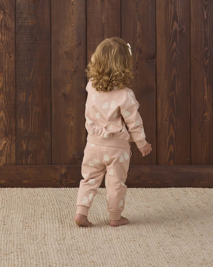 A young child with curly hair, wearing the quincy mae relaxed fleece sweatpant in sweet ghost—a peach-colored outfit with white patterns—stands on a textured beige rug, facing and touching a wooden wall.