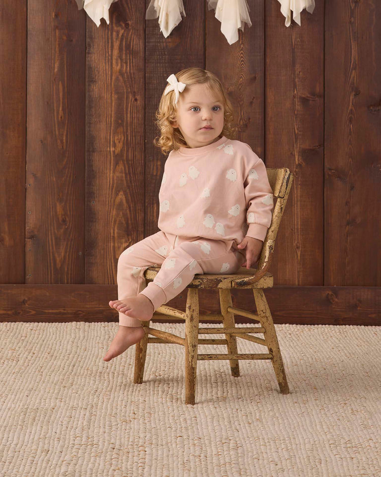 A young child with curly blonde hair and a bow sits barefoot on a wooden chair, wearing cozy quincy mae relaxed fleece sweatpants in the sweet ghost print and a light pink outfit with white patterns. The background features wood paneling, and the floor is covered with a textured beige rug.