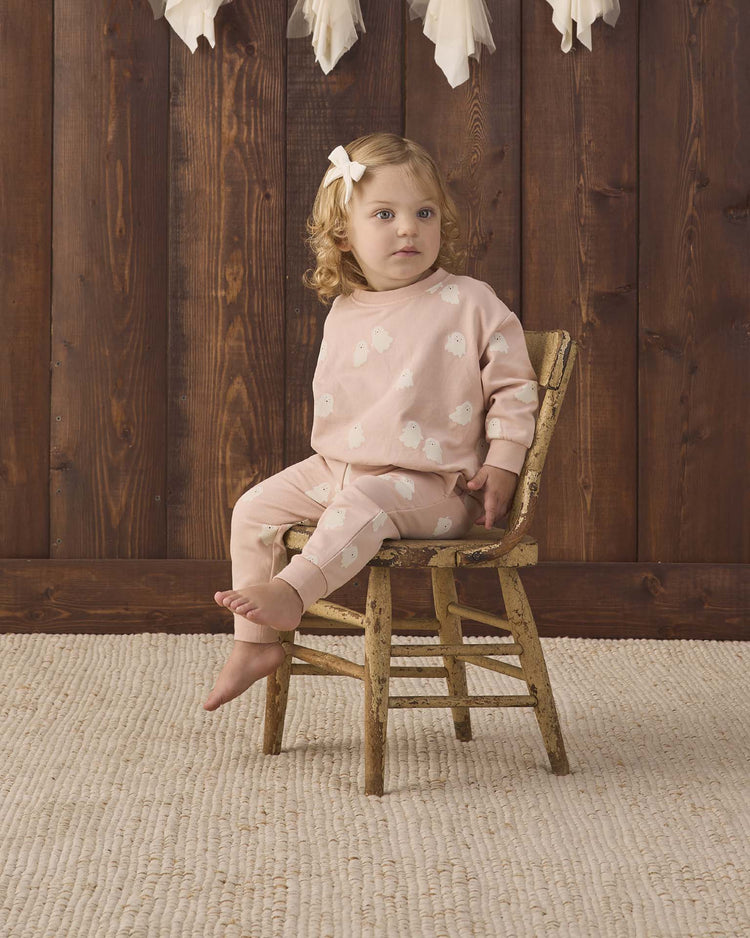 A young child with light brown hair, barefoot and wearing a white bow, sits on a rustic wooden chair. She is dressed in the quincy mae relaxed fleece sweatshirt in sweet ghost, featuring a charming ghost print and delicate white accents. The setting includes wooden paneling as the background and a textured cream rug on the floor.
