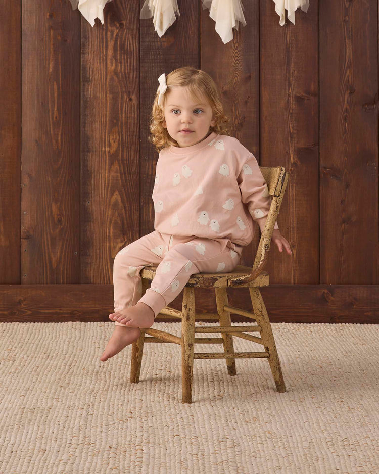 A young child with light brown hair sits barefoot on a rustic wooden chair, wearing quincy mae’s relaxed fleece sweatpant in sweet ghost paired with a pink outfit featuring white bird prints. The scene is set against a wooden wall and a textured beige rug.