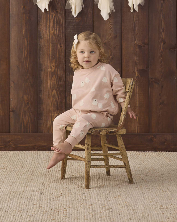 A young child with wavy blonde hair, wearing the quincy mae relaxed fleece sweatshirt in sweet ghost, sits barefoot on a rustic wooden chair against a dark wood wall and a beige textured rug.