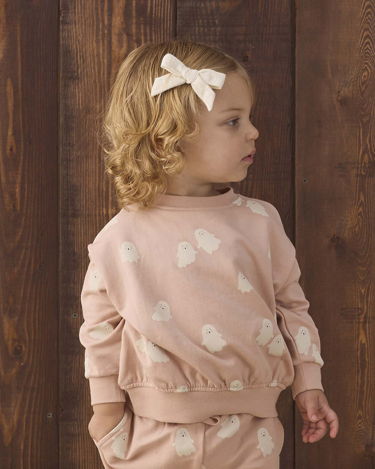 A young child with curly blond hair and a cream bow gazes to the side, wearing the Quincy Mae relaxed fleece sweatshirt in sweet ghost, standing in front of a wooden wall.