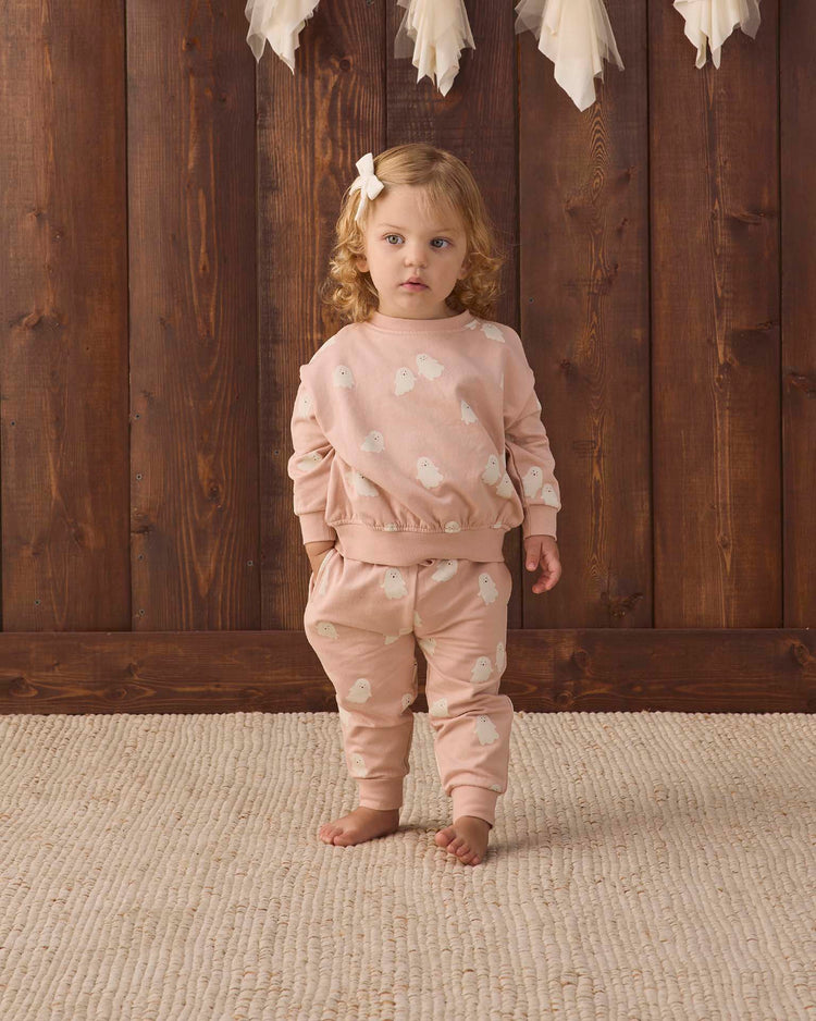 A young child with curly hair and a bow stands barefoot on a beige rug, wearing the quincy mae relaxed fleece sweatshirt in sweet ghost along with matching sweatpants, in front of a wooden wall adorned with white feather decorations above.