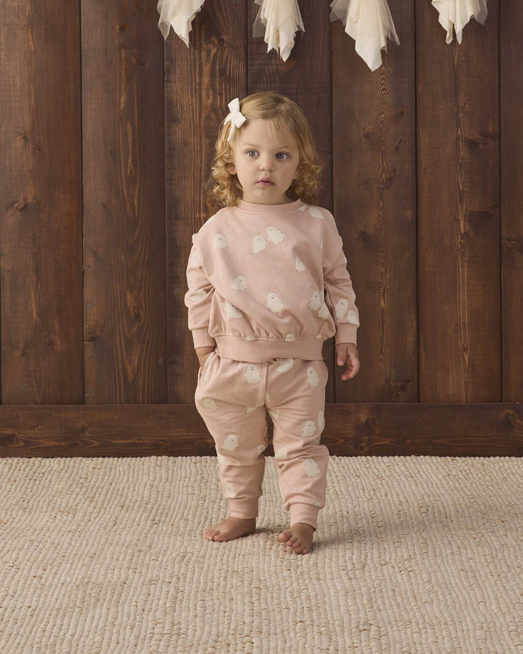 A young child with curly hair and a bow stands barefoot on a beige rug, wearing the quincy mae relaxed fleece sweatshirt in sweet ghost along with matching sweatpants, in front of a wooden wall adorned with white feather decorations above.