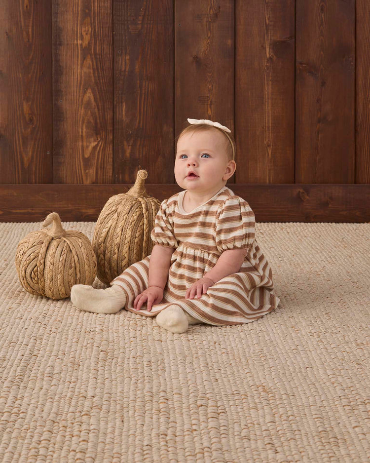 A baby wearing the quincy mae darla dress in spice stripe sits on a woven rug next to two decorative pumpkins, with a wooden wall in the background, accessorized with a matching headband.