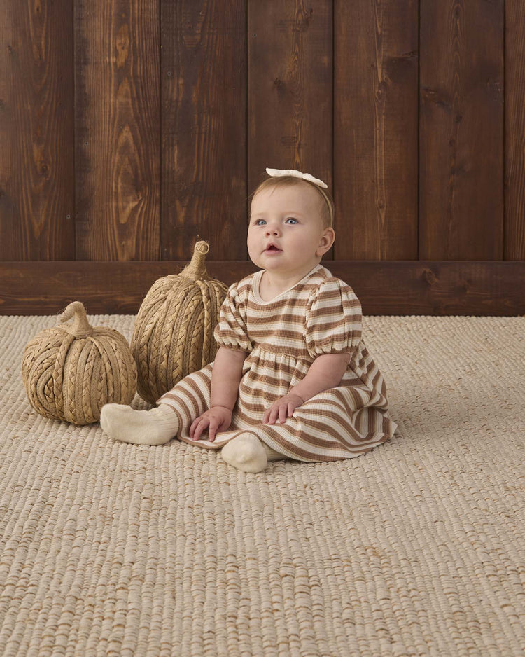 A baby wearing the quincy mae darla dress in spice stripe sits on a woven rug next to two decorative pumpkins, with a wooden wall in the background, accessorized with a matching headband.