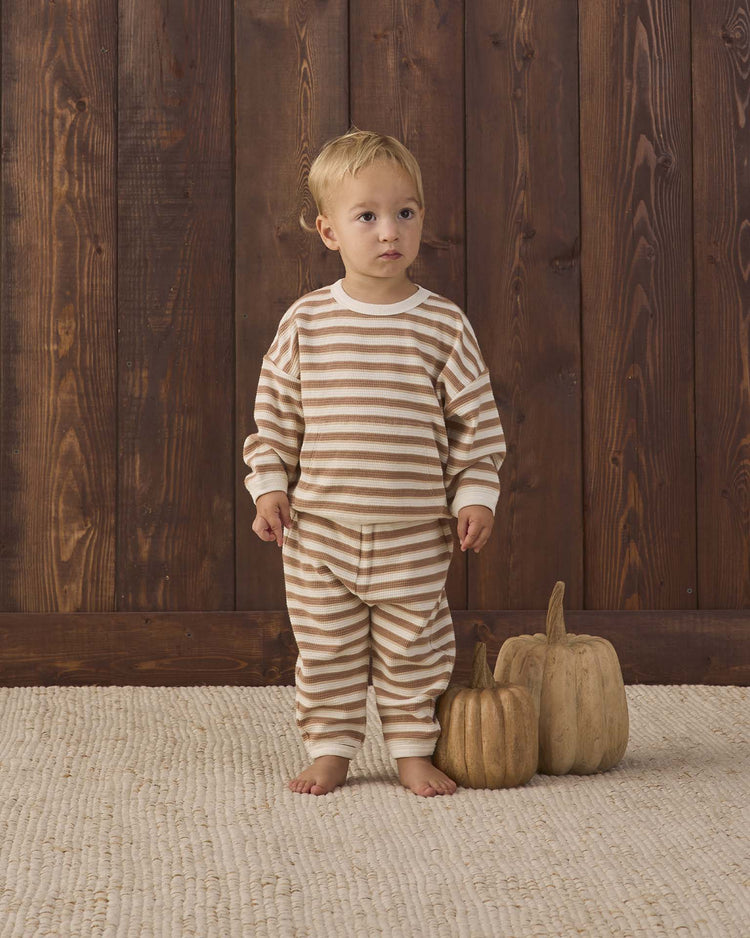 A toddler in the quincy mae waffle slouch set in spice stripe—organic cotton pajamas in beige and white stripes—stands barefoot on a textured rug beside two decorative pumpkins, with a wooden wall behind them.